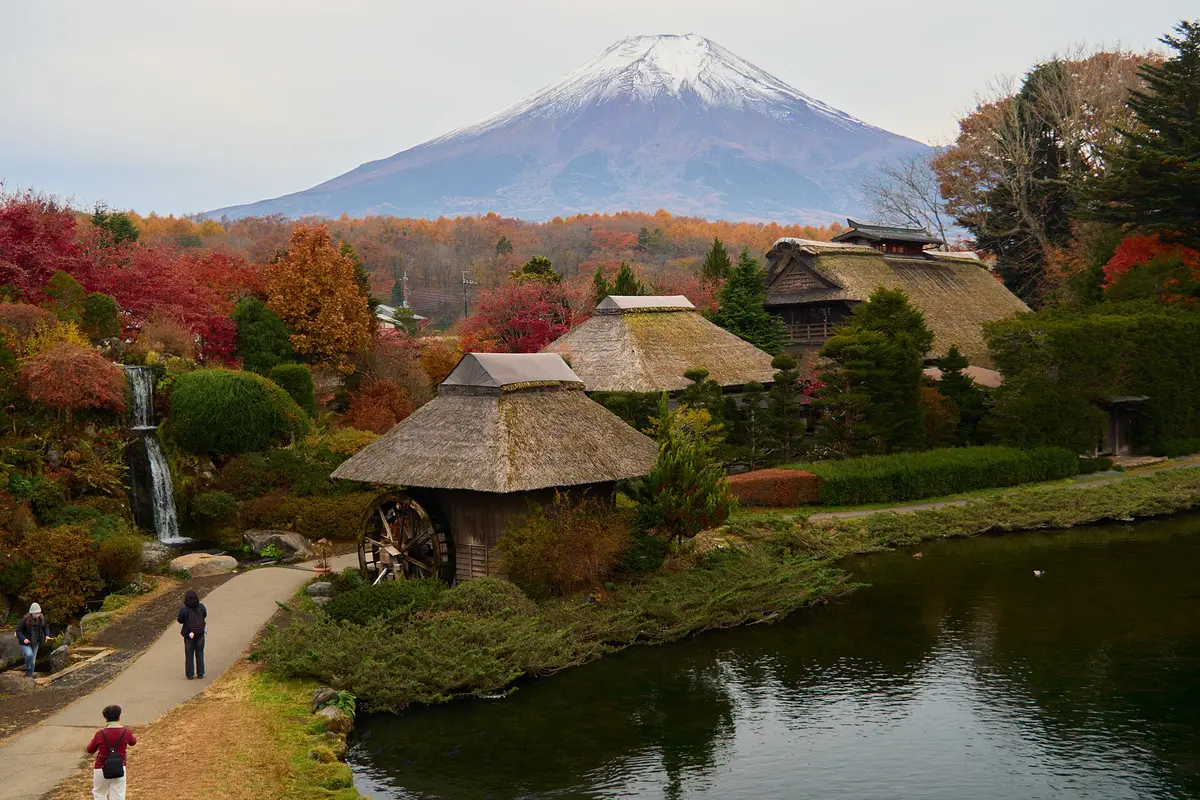 Thatched buildings and a waterwheel beside a pond at Oshino Hakkai in autumn, with snow-capped Mount Fuji behind